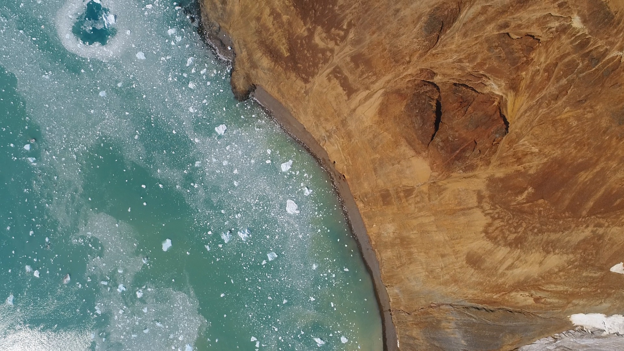 Aerial view of a turquoise lagoon meeting dark volcanic rock on the Antarctic coastline